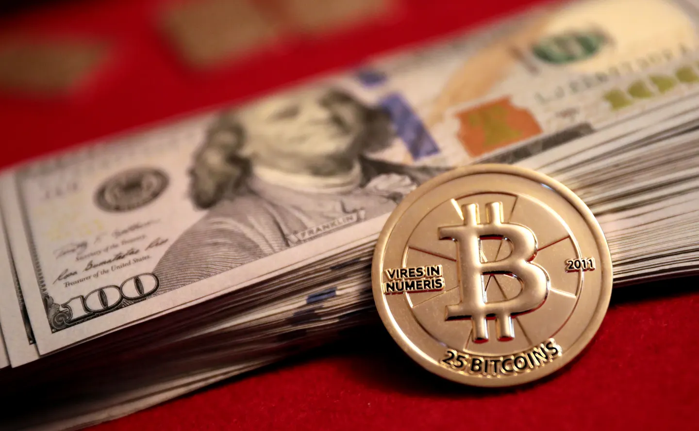 A gold Casascius Bitcoin collectible leaning against a stack of U.S. hundred-dollar bills