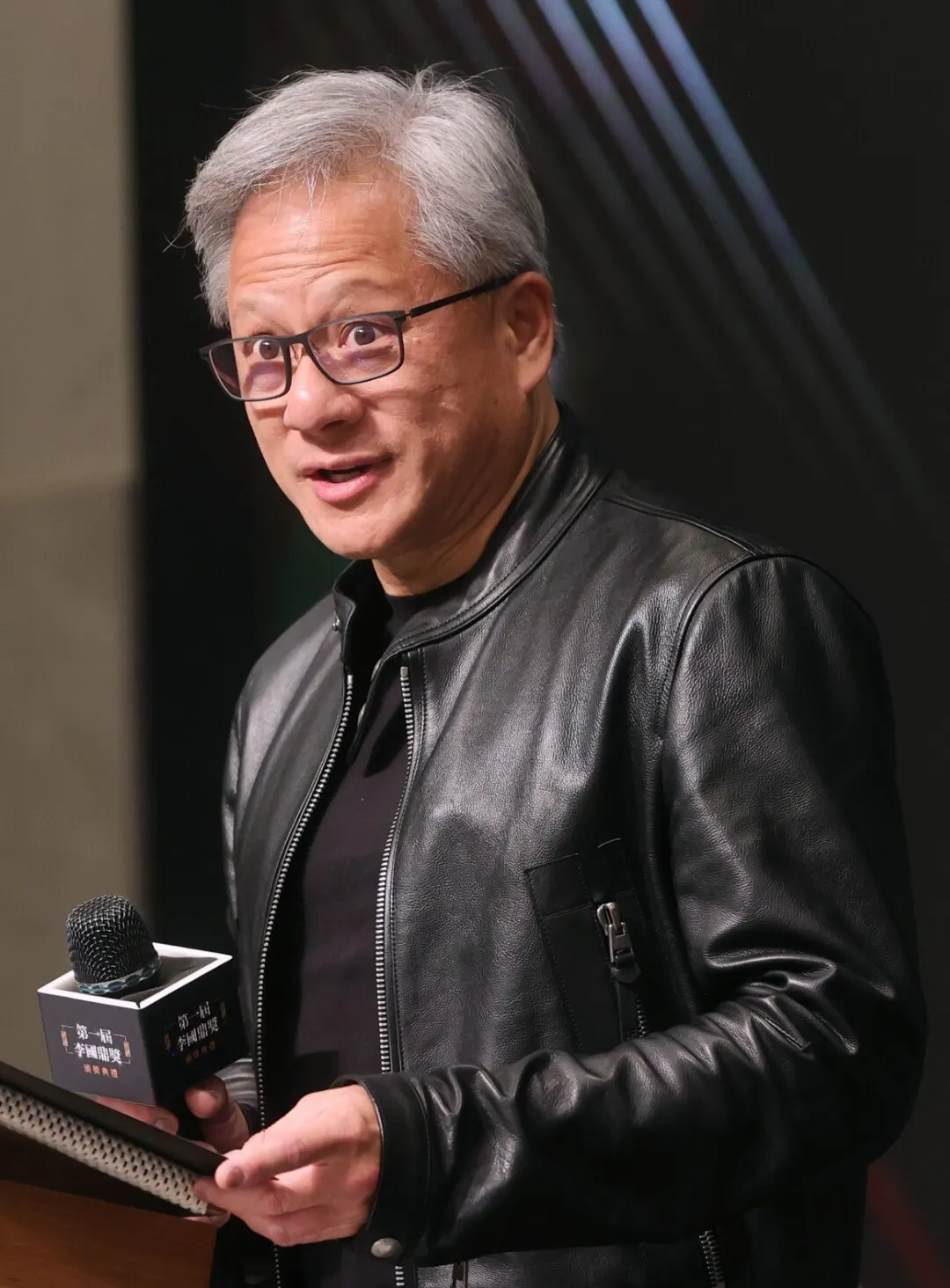 Jensen Huang wearing glasses and a black leather jacket, holding a microphone