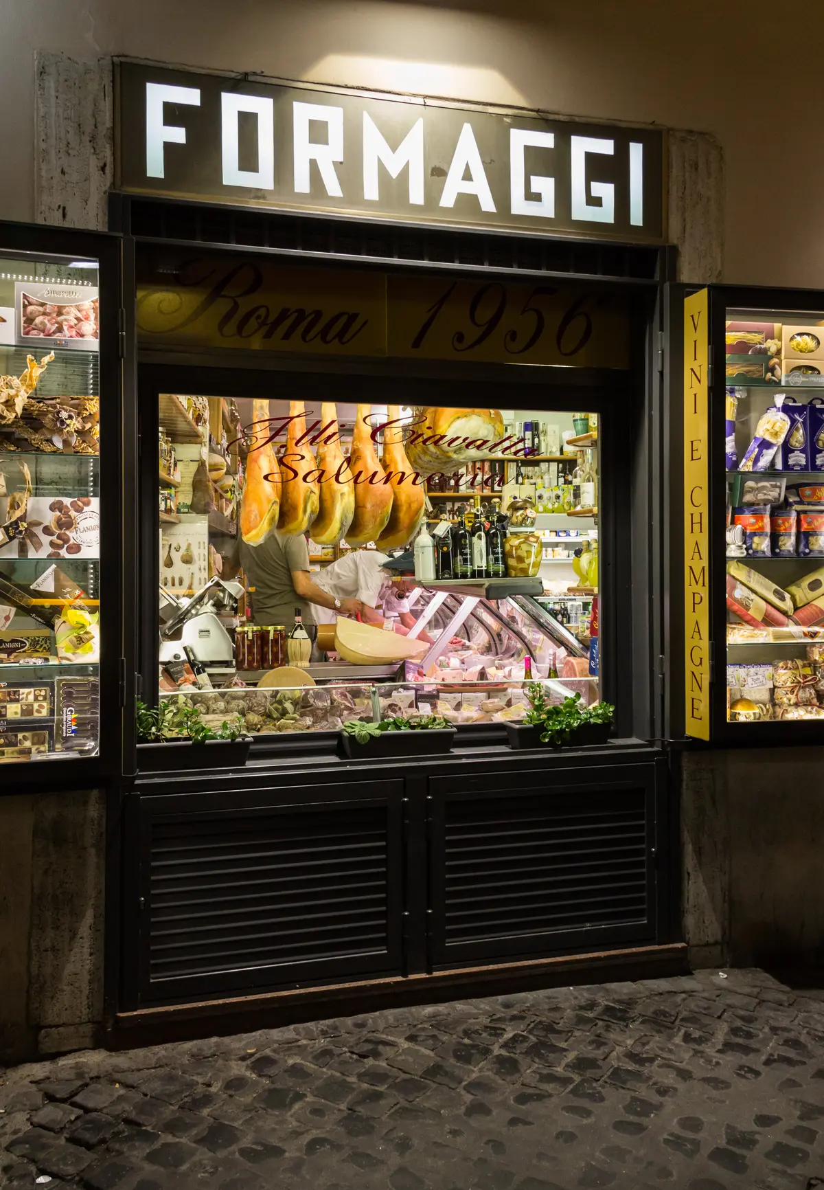 Vetrina illuminata di una salumeria di Roma con formaggi, salumi e due addetti dietro il banco
