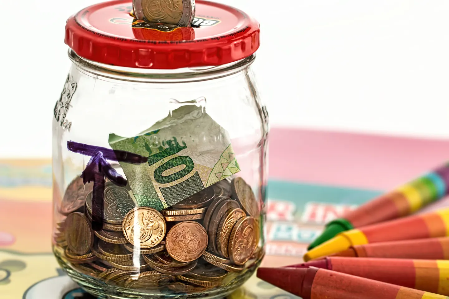 A glass savings jar containing coins and a folded banknote beside colored pencils